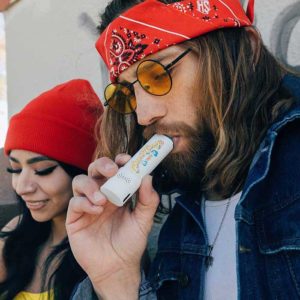 Man with a red bandanna and blue jean jacket smoking a Cheech and Chong Mambo vaporizer for cannabis flower. There is a woman with a red hat and a yellow shirt sitting next to the man.