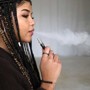 A woman with long braids and a black sweater smoking a VPM glass cart with cannabis oil on a black oil pen.